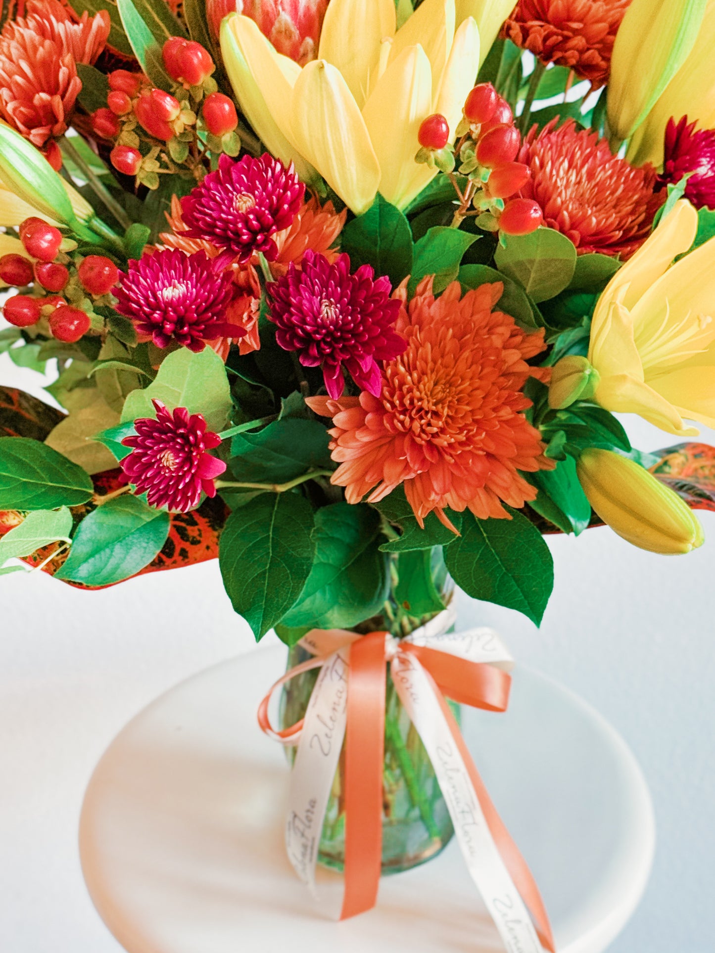 Bouquet of colorful flowers in a clear vase with an orange ribbon on a white surface.