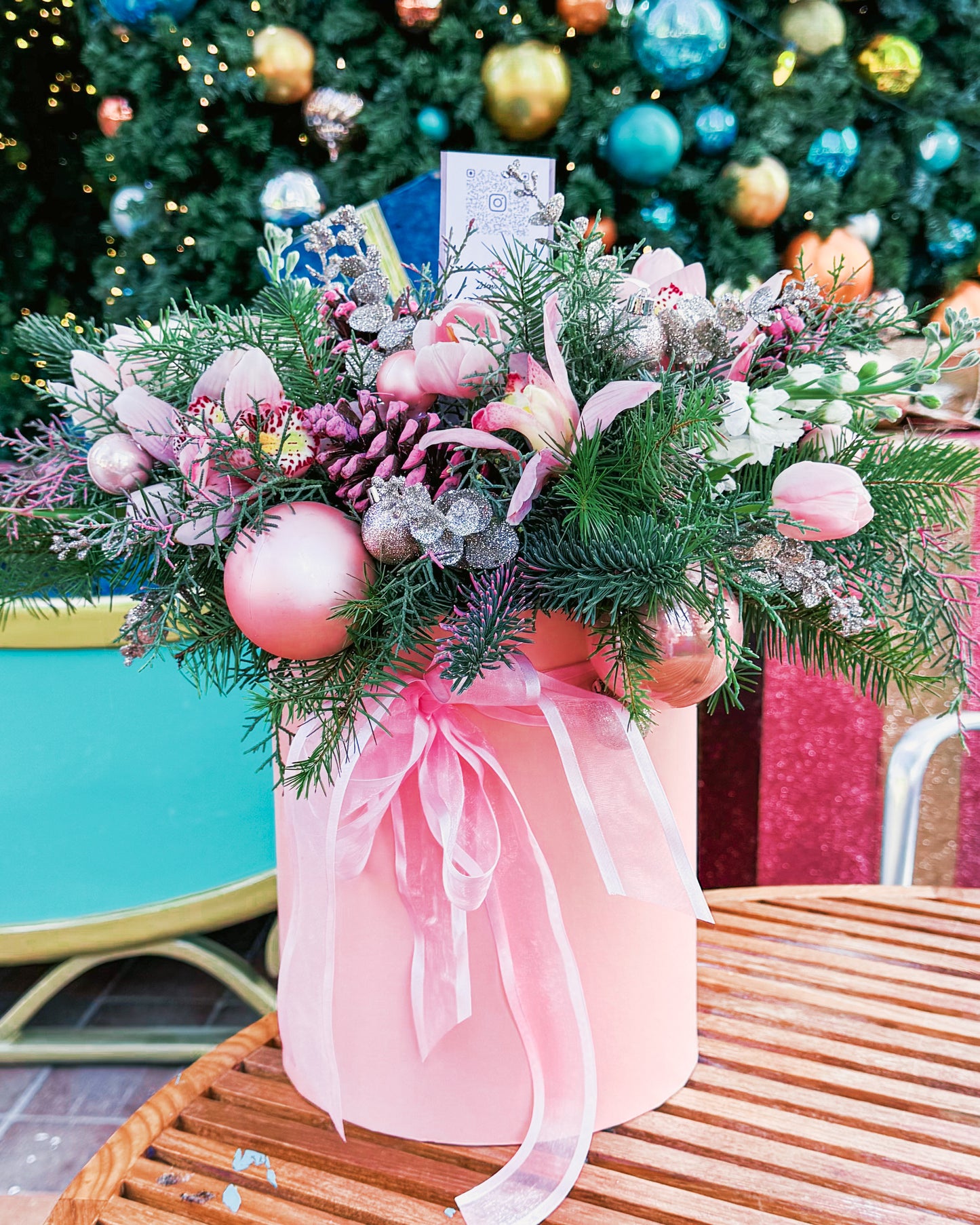 Decorative floral arrangement with pink ornaments and greenery on a table in front of a Christmas tree.