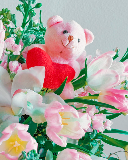 Pink teddy bear with a red heart surrounded by pink and white flowers on a light gray background