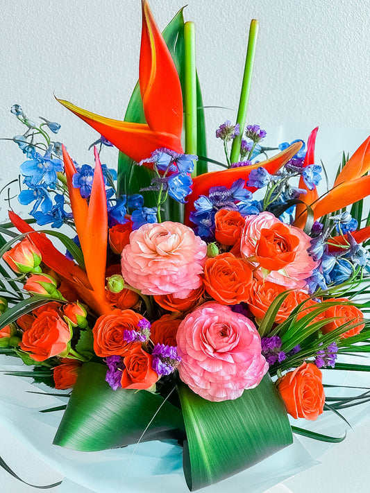 Spice Bloom by Zelenaflora, a tropical spring bouquet with orange spray roses, pink ranunculus, blue delphinium, purple statice, and sculptural greenery in Los Angeles