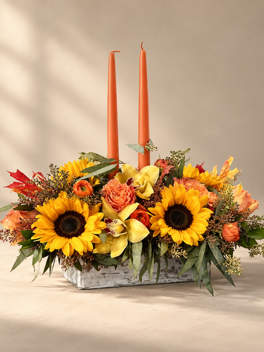 Autumn centerpiece with two unlit peach candles, sunflowers, yellow orchids, orange roses and ranunculus in a low rectangular container