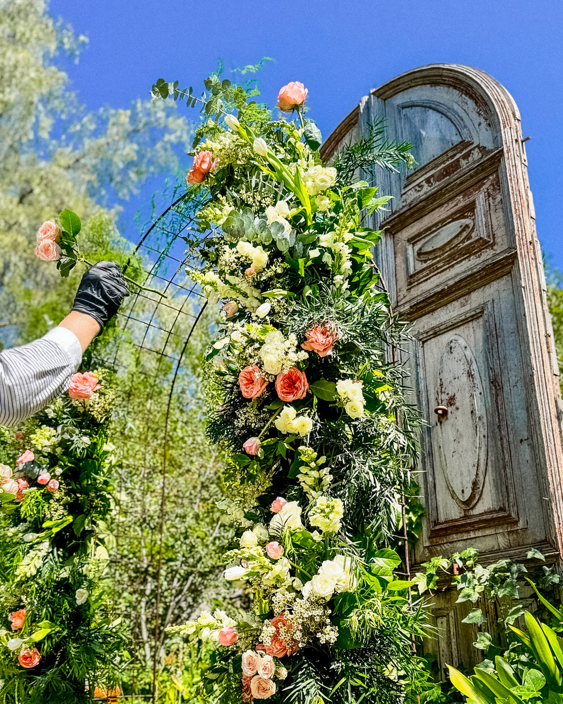 Floral archway being adjusted against a rustic wooden door with trees and blue sky in the background