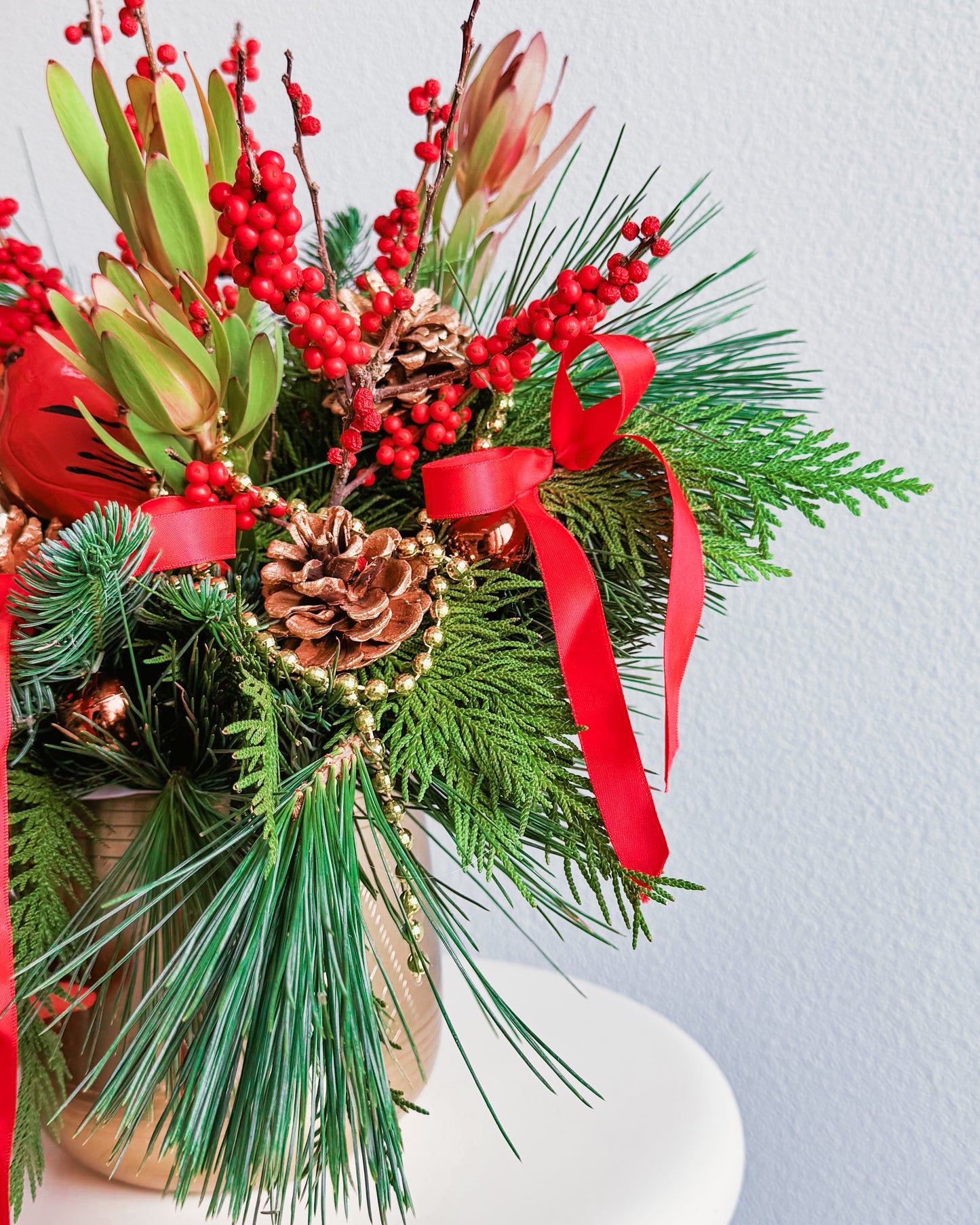 Christmas-themed floral arrangement with red berries, pine cones, and ribbons on a light background