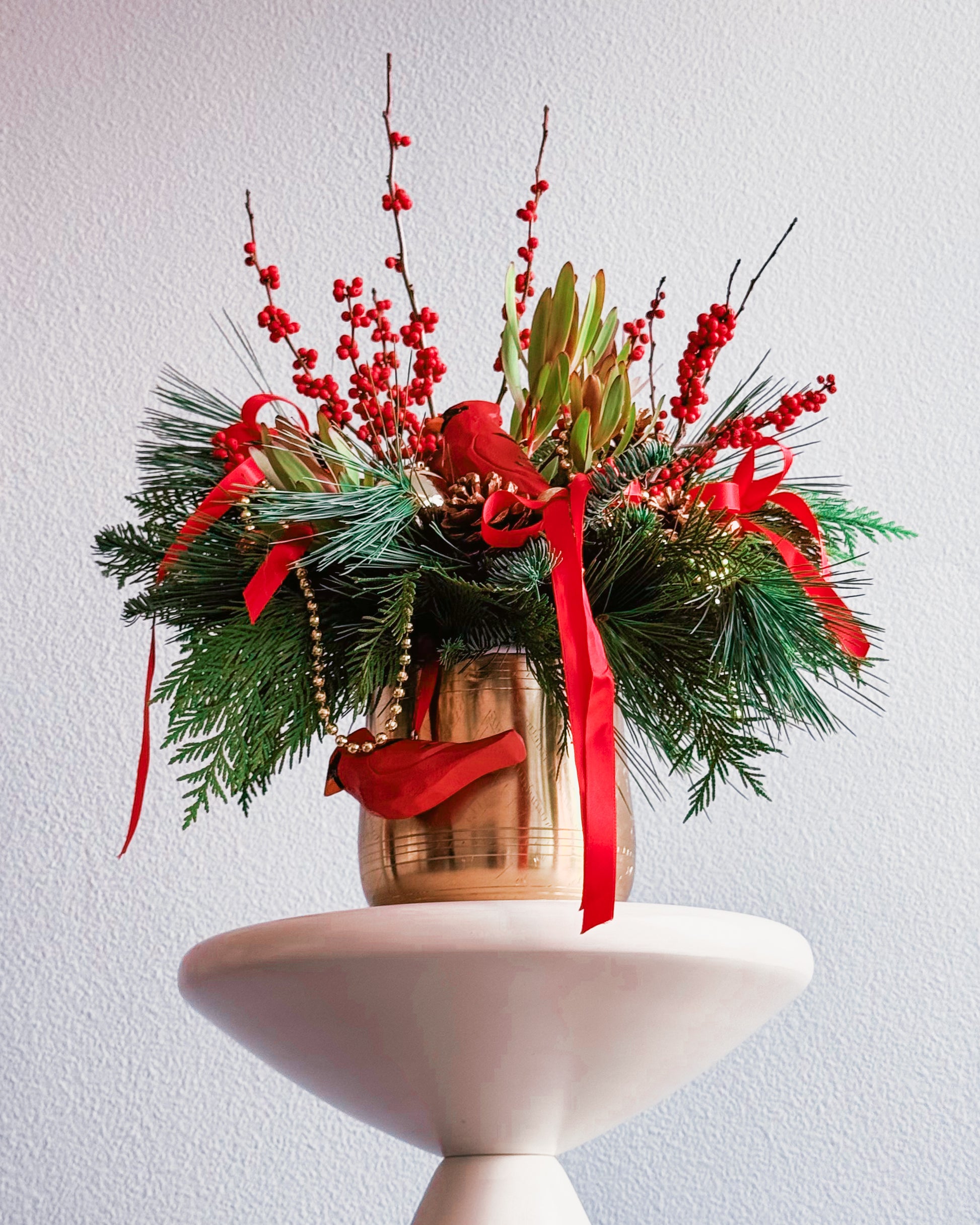 Decorative floral arrangement with red ribbons and berries on a white pedestal against a light gray background