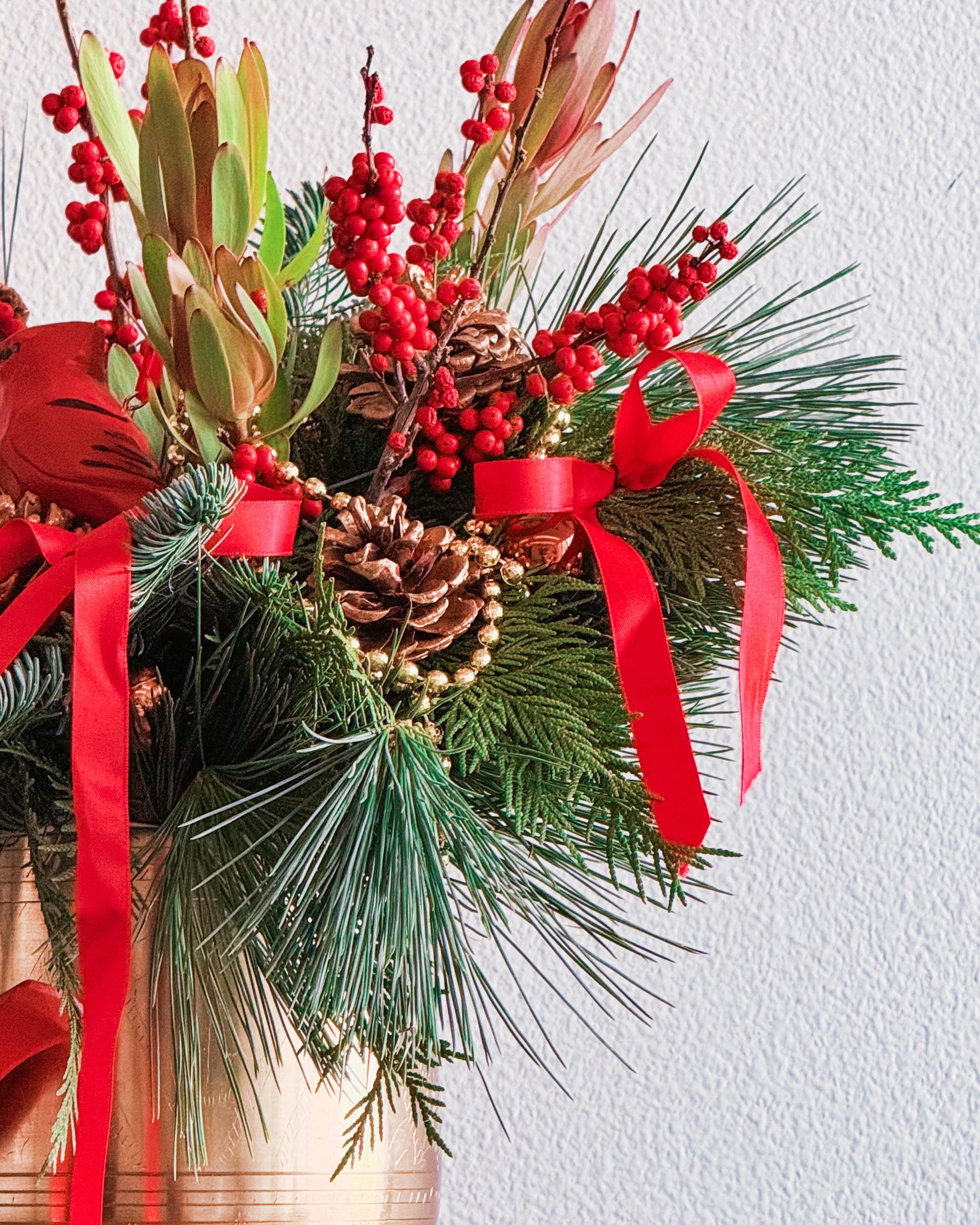 Christmas-themed floral arrangement with red ribbons, pine cones, and berries on a white background
