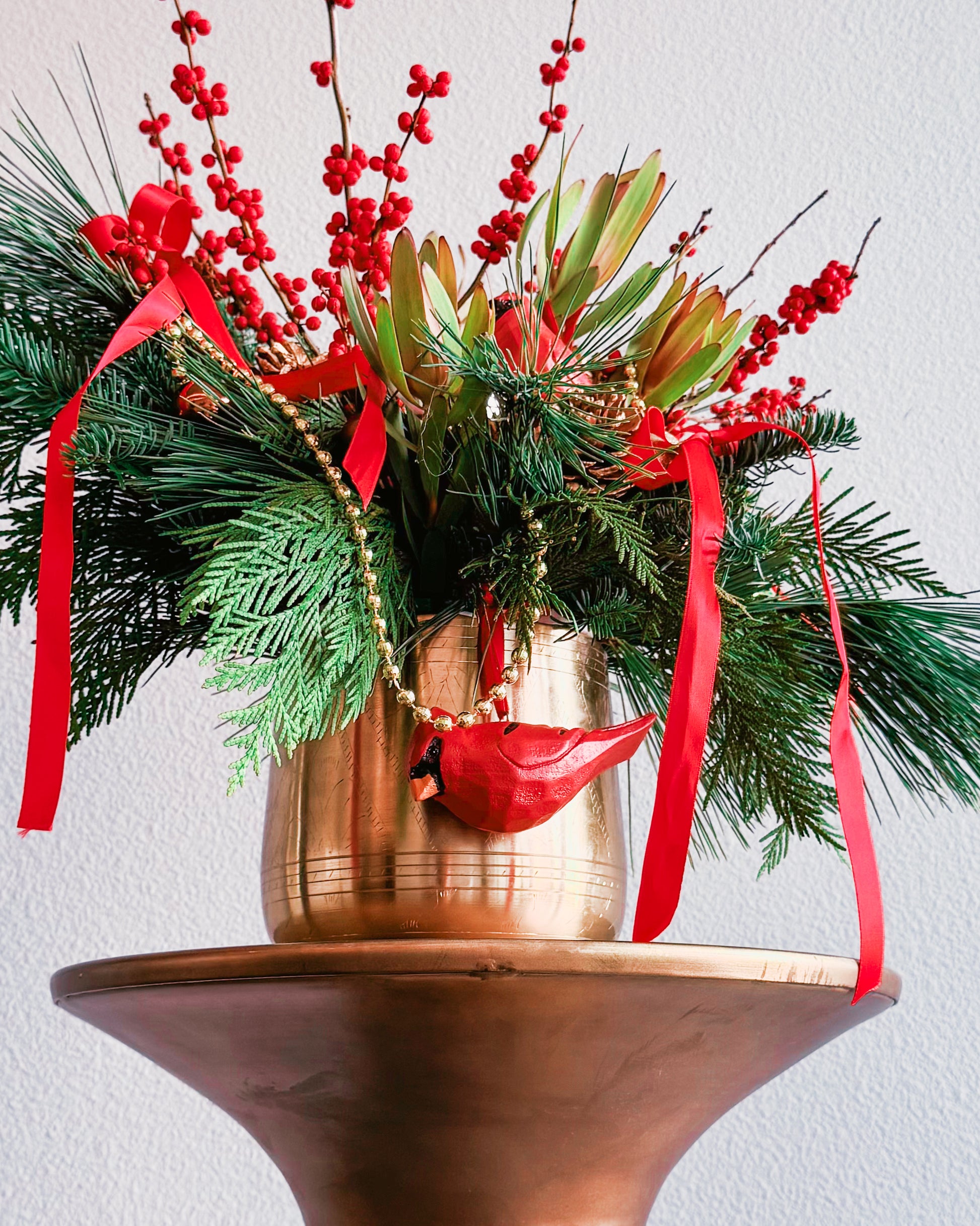 Decorative Christmas arrangement with greenery, red berries, and ribbons in a gold pot on a white background.