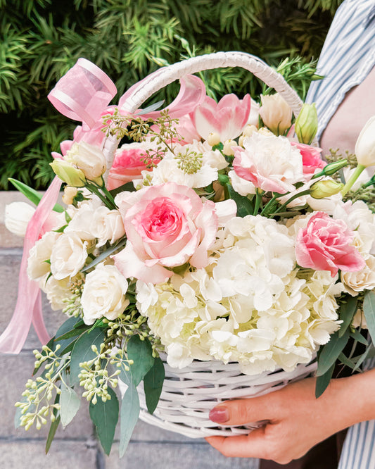 Shades of Caress romantic basket bouquet with pink roses, creamy white spray roses, pink tulips, white hydrangeas, lisianthus buds, eucalyptus, and pink ribbon by Zelena Flora