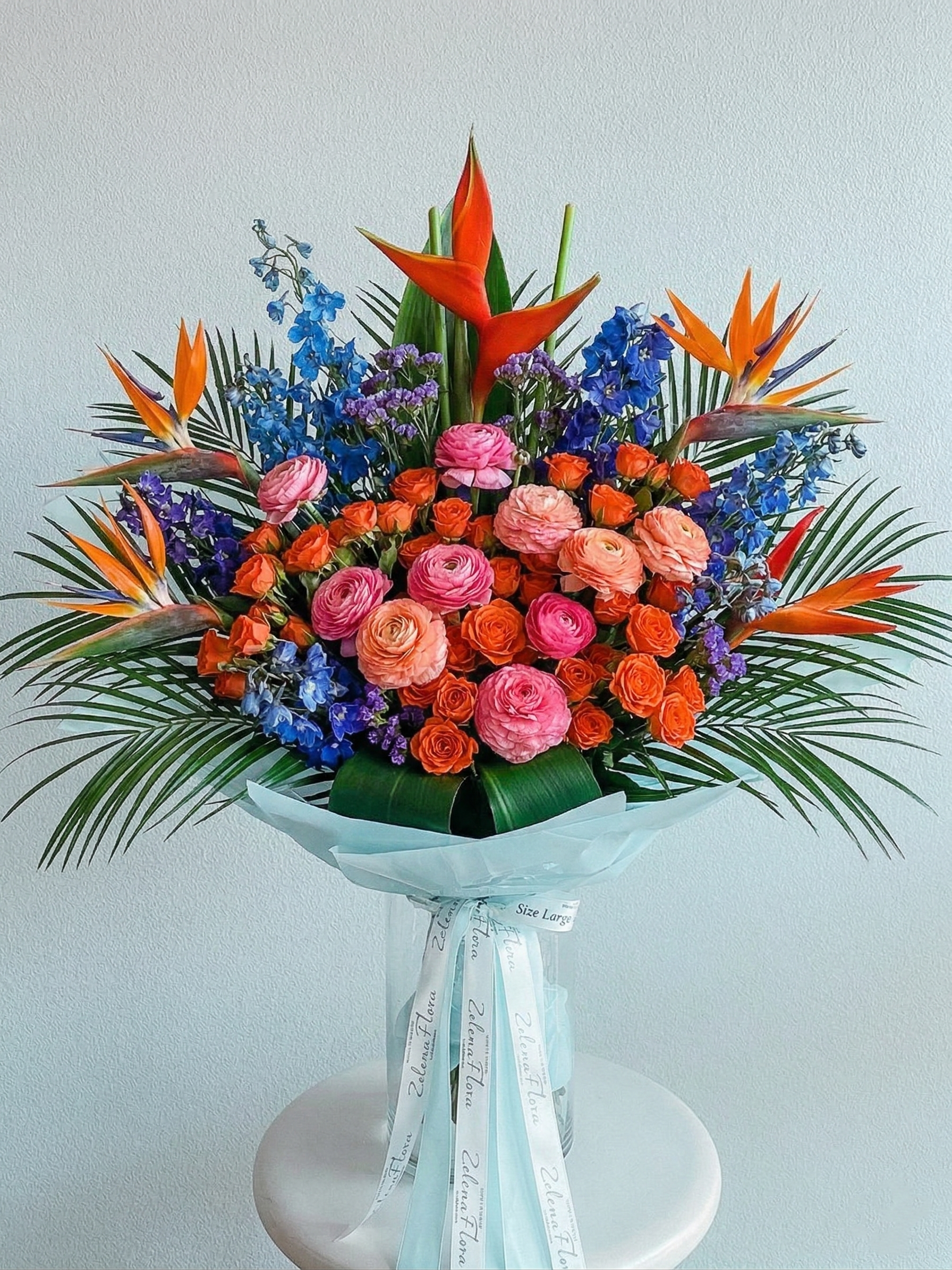 Spice Bloom by Zelenaflora, a tropical spring bouquet with orange spray roses, pink ranunculus, blue delphinium, purple statice, and sculptural greenery in Los Angeles