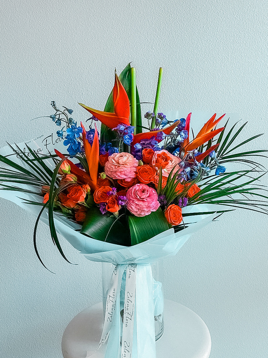 pice Bloom by Zelenaflora, a tropical spring bouquet with orange spray roses, pink ranunculus, blue delphinium, purple statice, and sculptural greenery in Los Angeles