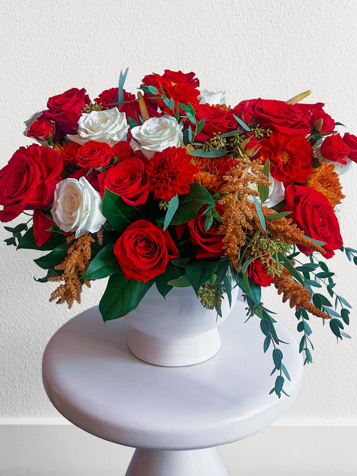 Full view of Crimson Charm flowers in a white pedestal vase, with red and white blooms, golden accents and cascading greenery.