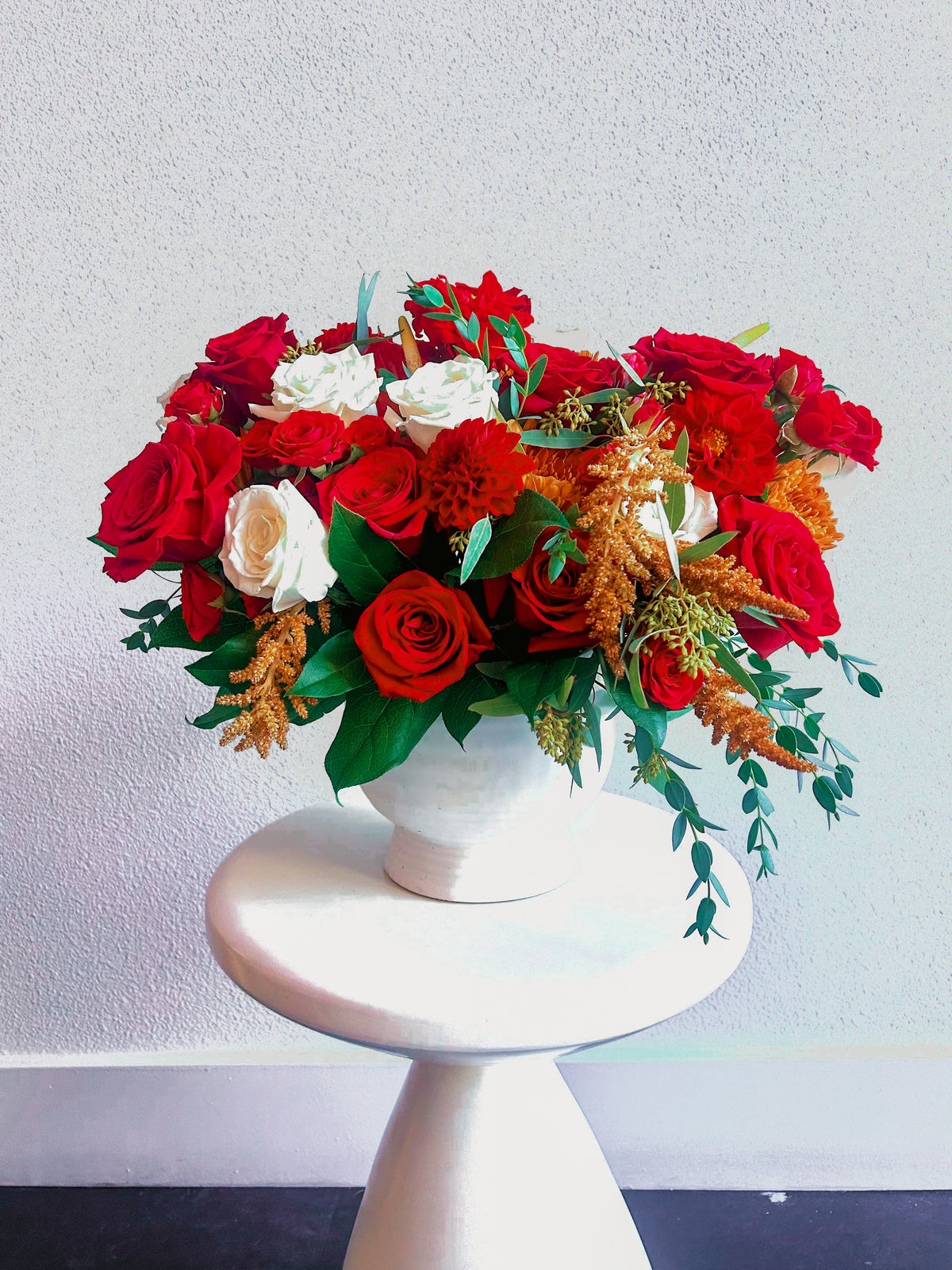 Crimson Charm vase arrangement with red and white roses, red dahlias, copper chrysanthemums and trailing greenery in a white pedestal vase.