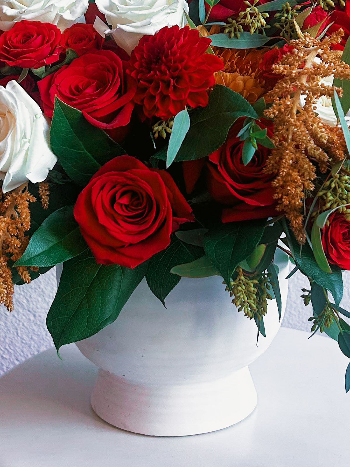 Close view of red roses and a red dahlia above the white pedestal vase in Zelena Flora’s Crimson Charm arrangement.