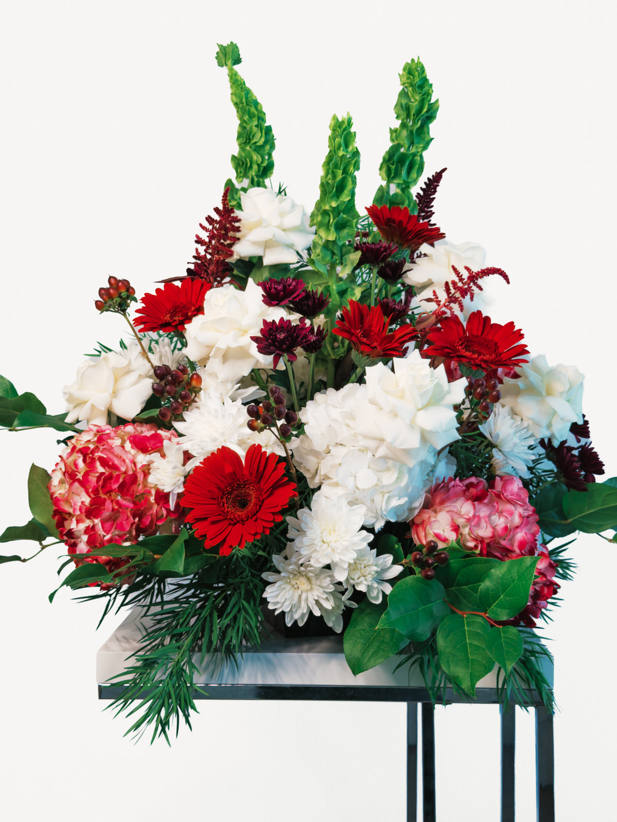Floral Fanfare wedding and event arrangement with red gerberas, white roses, pink hydrangeas and tall green Bells of Ireland; stand shown for styling.