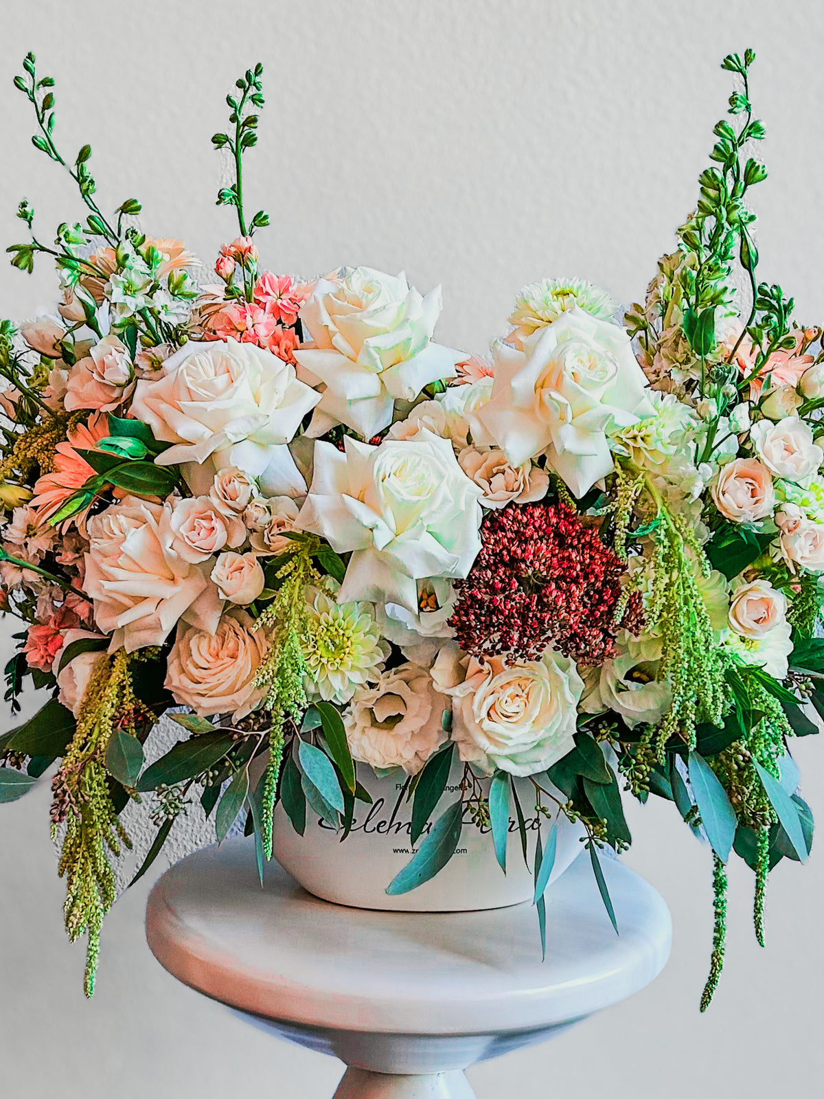 Closer view of Gentle Gesture with ivory and champagne roses, peach-toned flowers and green accents in a rounded white Zelena Flora vase.