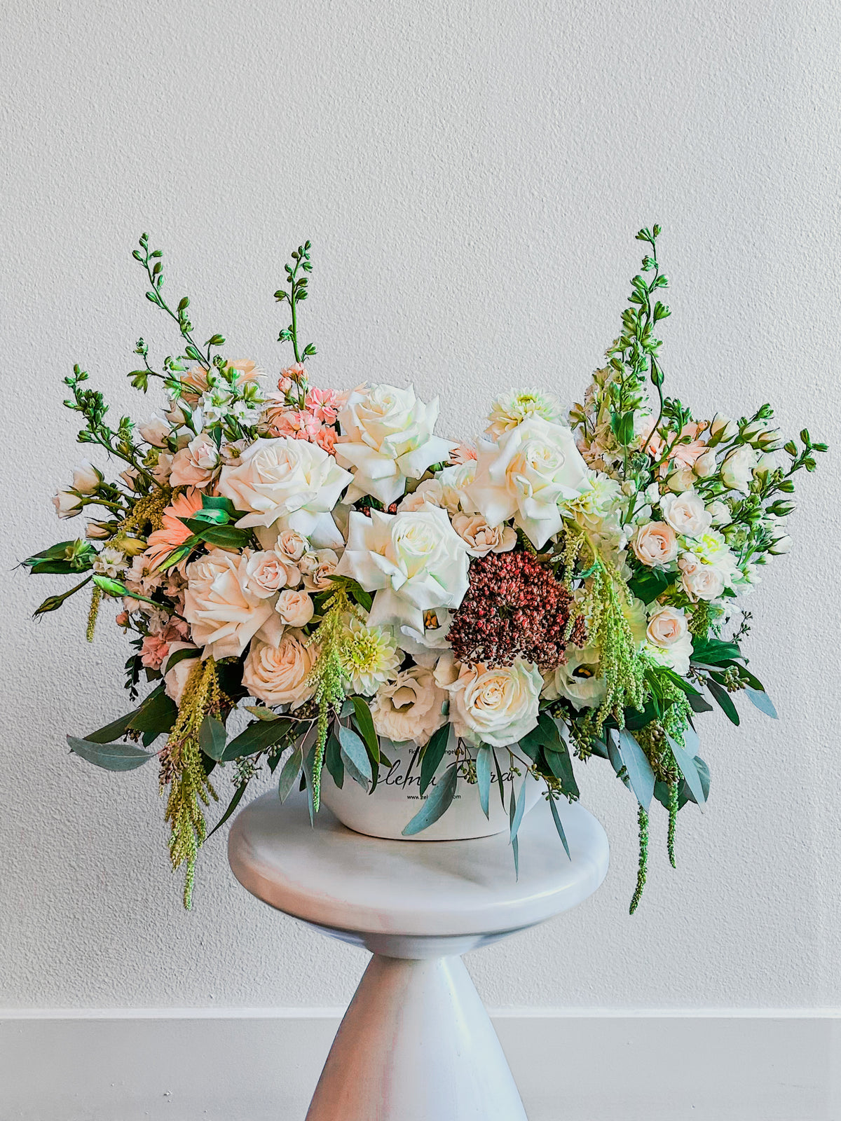 Gentle Gesture vase arrangement with ivory roses, cream spray roses, soft peach blooms, burgundy accents and trailing eucalyptus on a separate display stand.