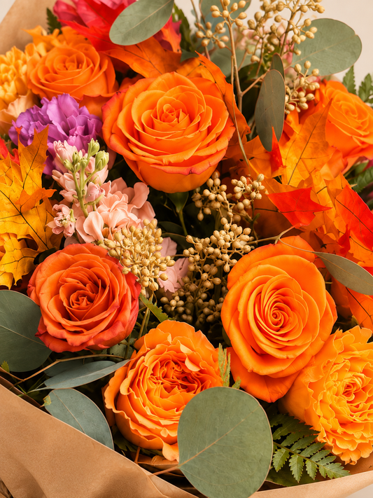Close-up of Golden Harvest orange roses, golden chrysanthemum, peach and lilac stock, eucalyptus and preserved fall leaves in kraft wrap